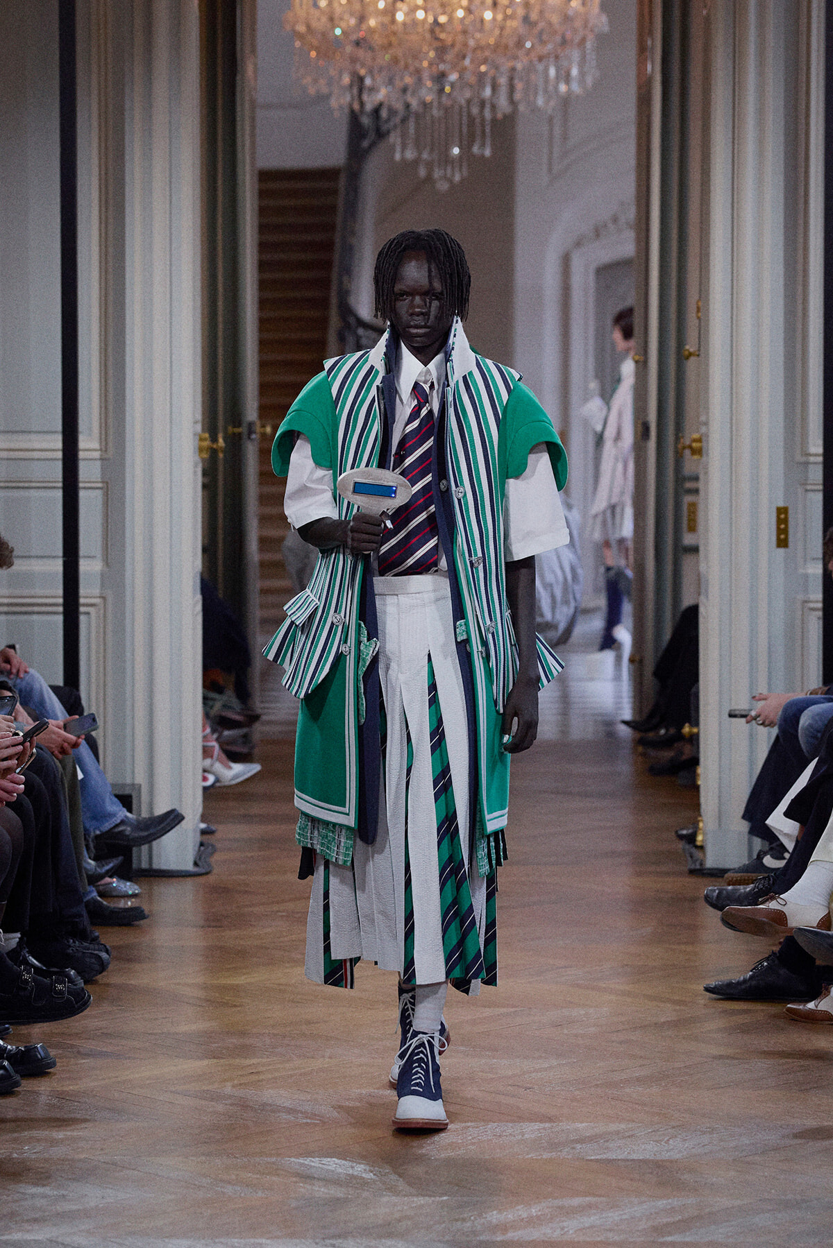 Model walks the runway wearing a green and striped layered ensemble with a white shirt and tie, pleated skirt, in a grand hall with a chandelier above.