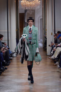 Female model on a runway wearing a green plaid suit with white shirt and striped sweater, carrying a matching green plaid handbag and striped coat atop her arm.