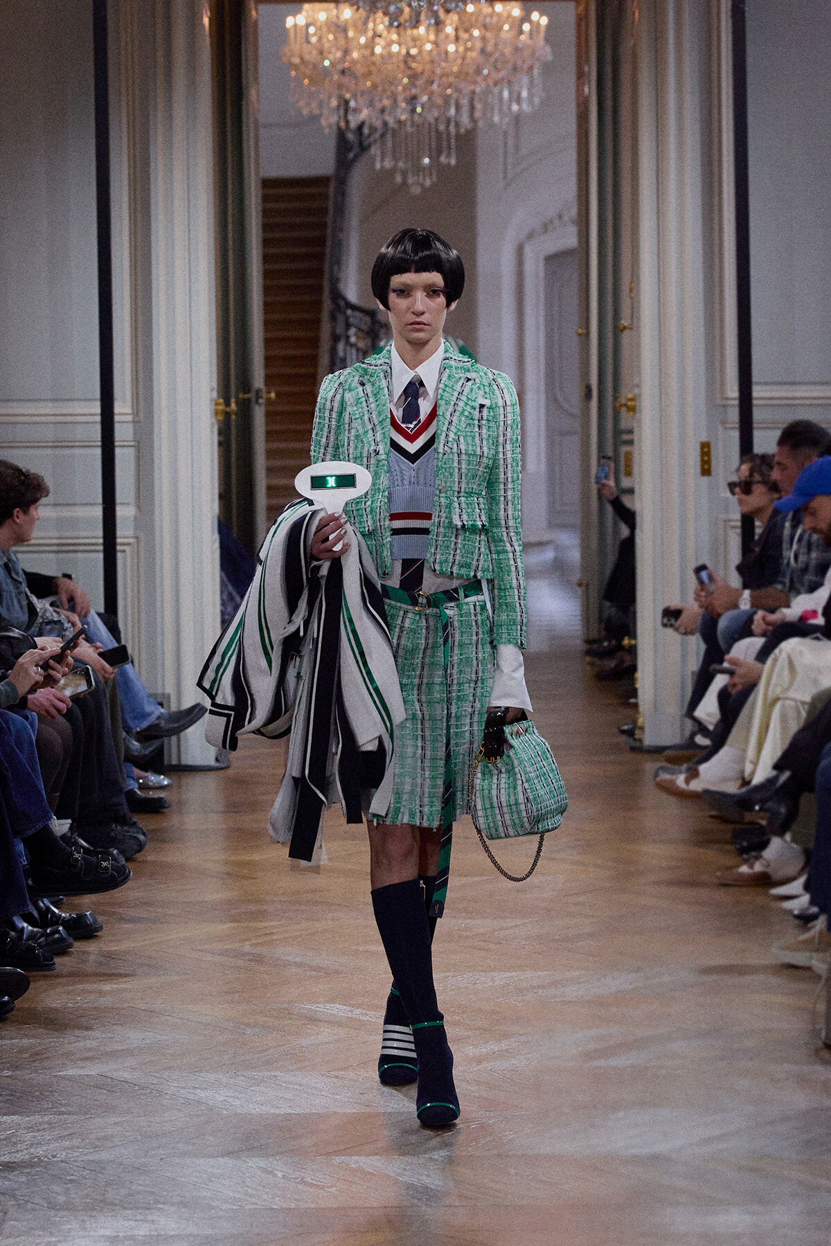 Female model on a runway wearing a green plaid suit with white shirt and striped sweater, carrying a matching green plaid handbag and striped coat atop her arm.