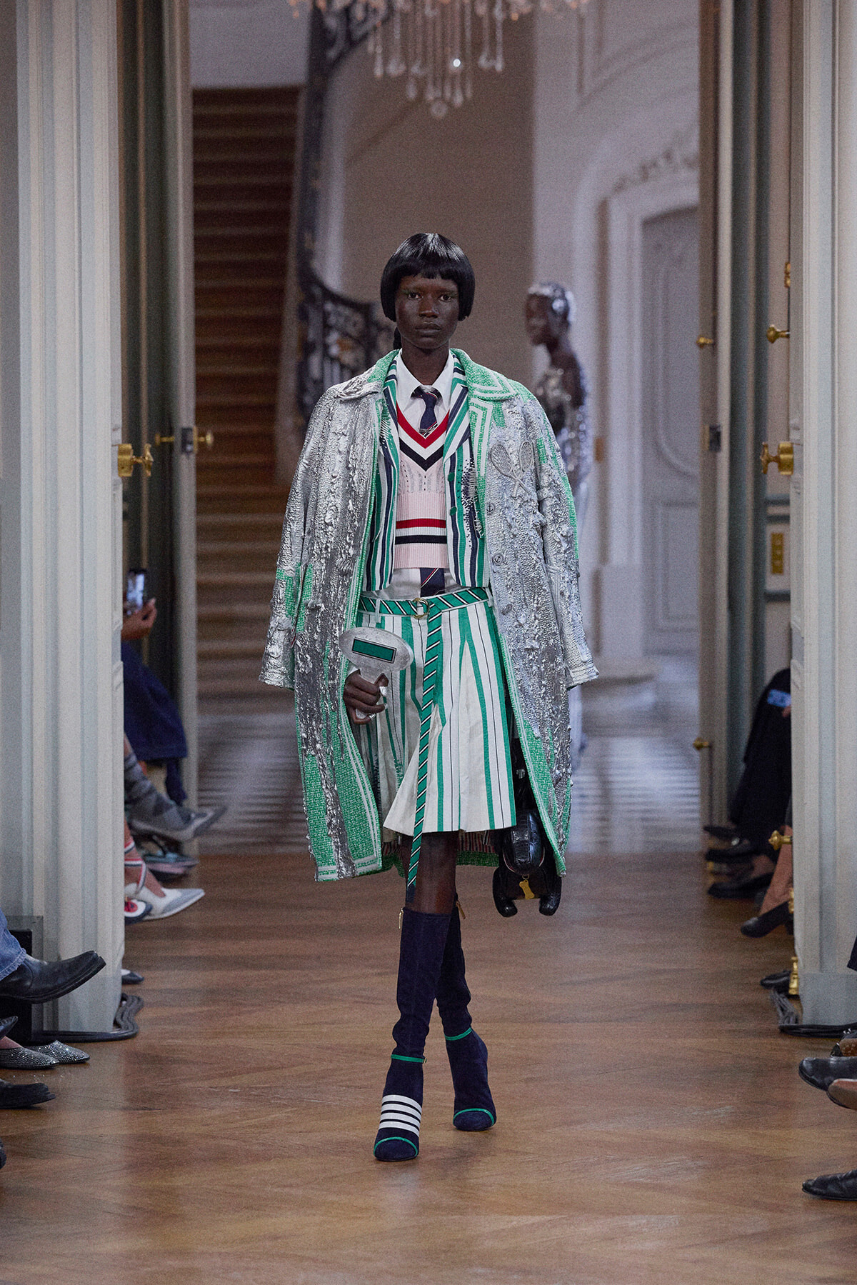 Model walks a runway in a metallic silver-and-green striped coat over a color-blocked knit top and skirt, paired with navy boots in a grand hallway.