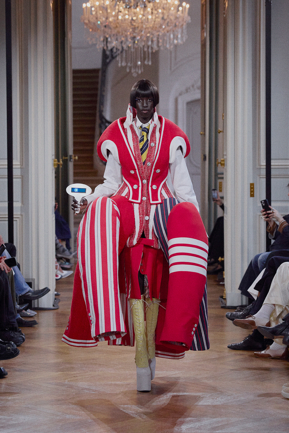 Model walks a runway in a bold red and white sculptural coat with oversized striped leg extensions, chandeliers overhead in a grand room.