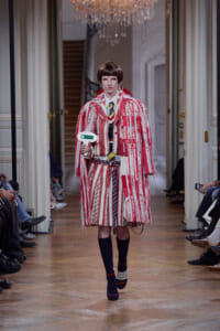 Runway model wearing a bold red-and-white fringed coat over a striped suit, holding a small white handheld prop, in a chandelier-lit venue.