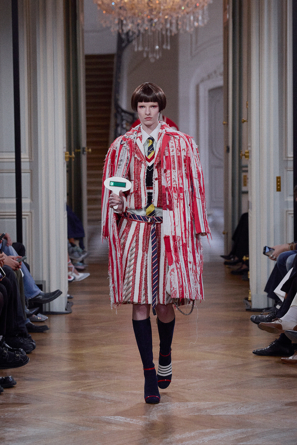 Runway model wearing a bold red-and-white fringed coat over a striped suit, holding a small white handheld prop, in a chandelier-lit venue.