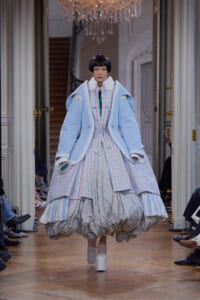 Model walking a runway in an oversized light-blue coat with plaid inner layers and a voluminous striped skirt, white platform boots.