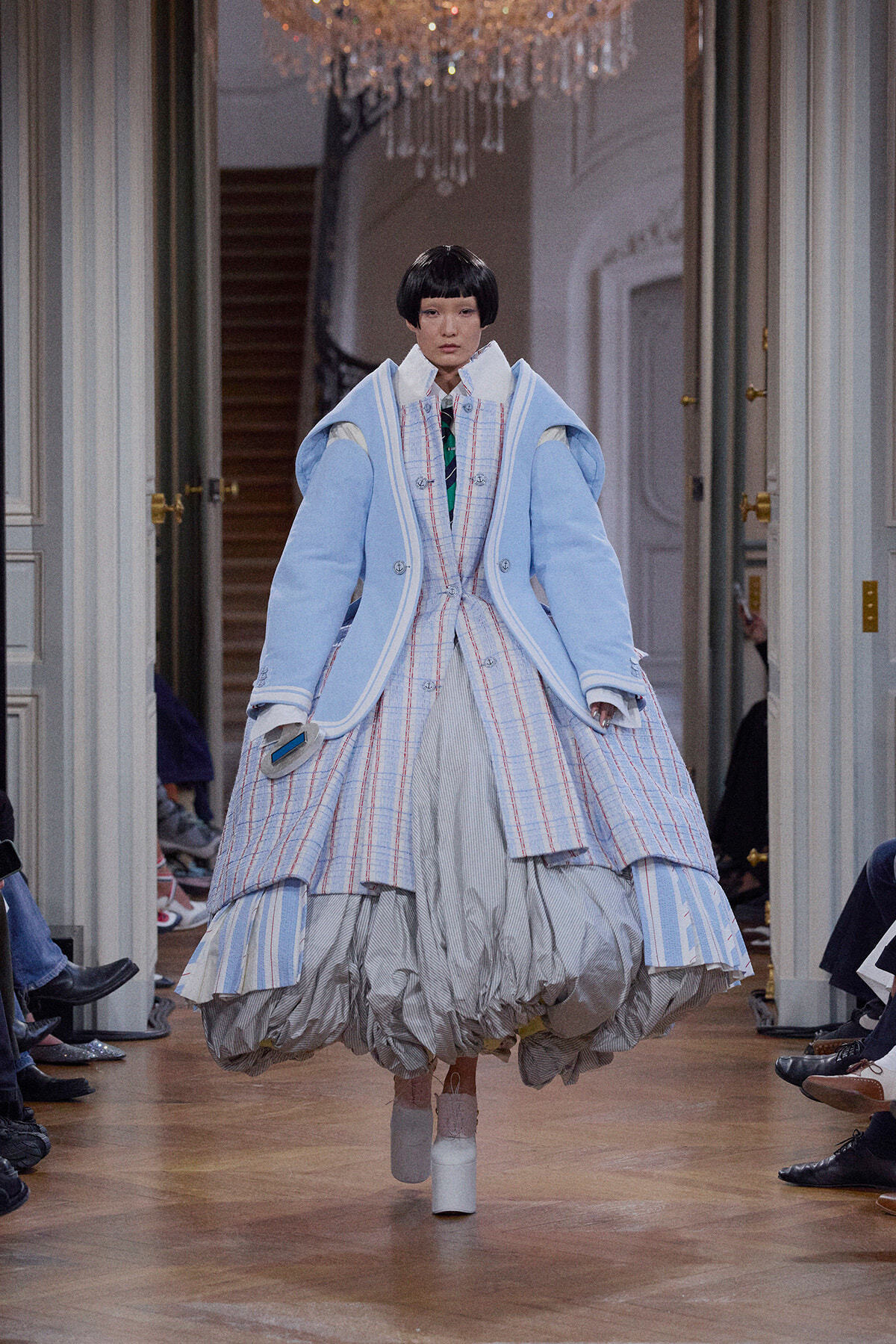 Model walking a runway in an oversized light-blue coat with plaid inner layers and a voluminous striped skirt, white platform boots.
