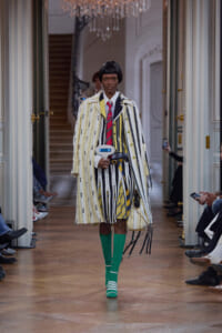 Runway model in a bold striped coat and matching striped suit with a red tie and bright green socks walking a chic hallway runway.