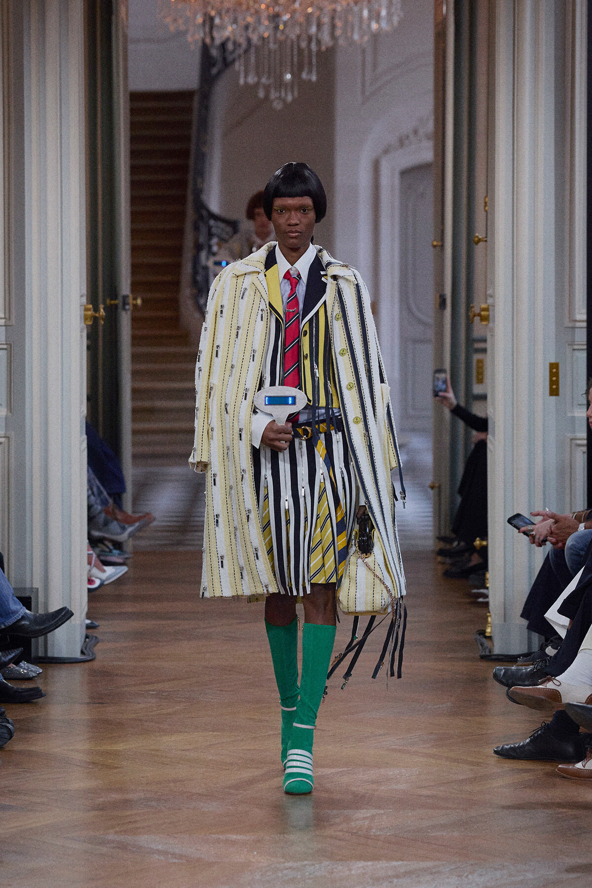 Runway model in a bold striped coat and matching striped suit with a red tie and bright green socks walking a chic hallway runway.