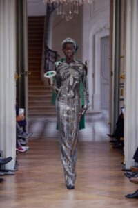 Black model walks a runway in a full-length silver sequined jumpsuit with geometric patterns, holding a white, microphone-like prop with a green light edge.