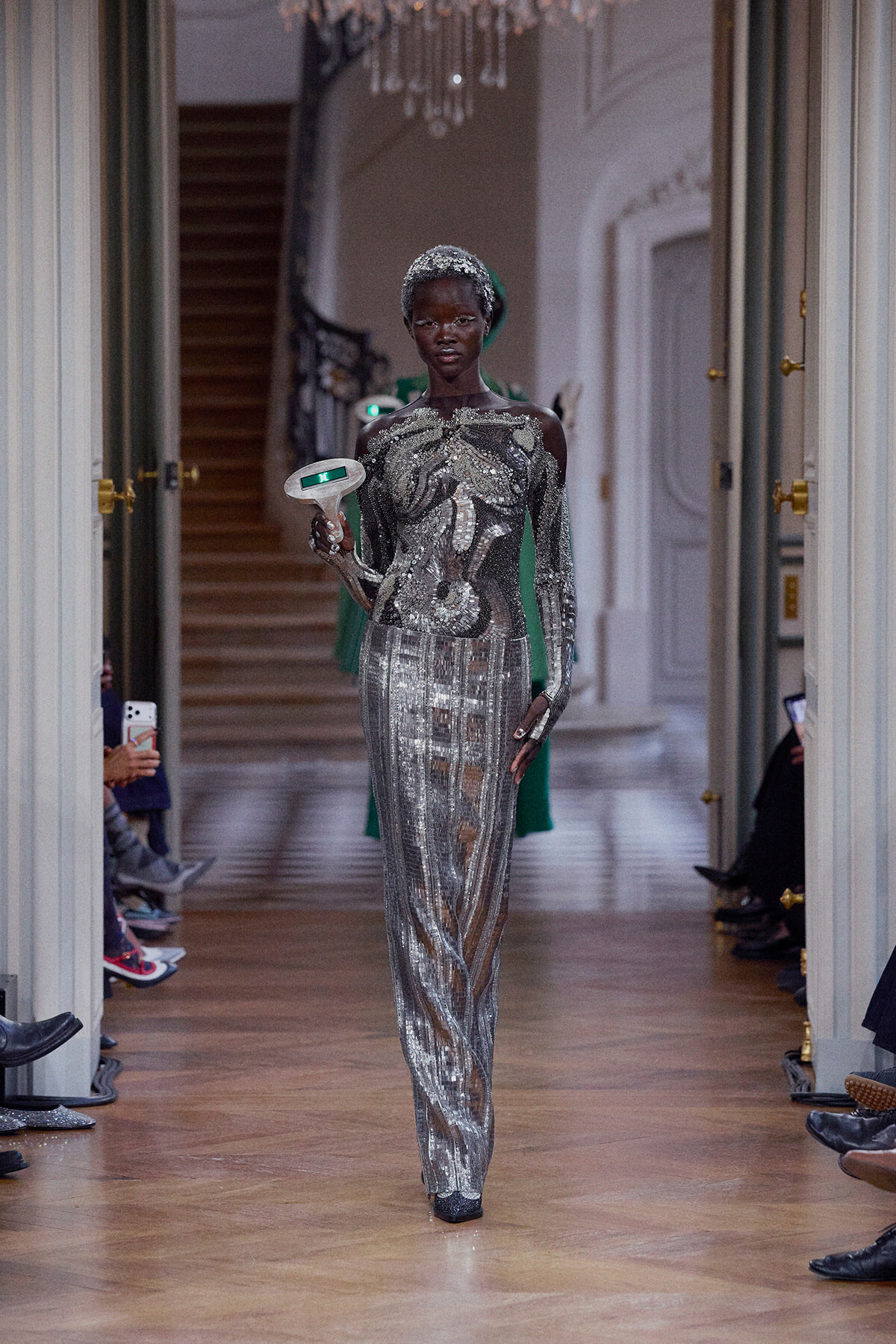 Black model walks a runway in a full-length silver sequined jumpsuit with geometric patterns, holding a white, microphone-like prop with a green light edge.