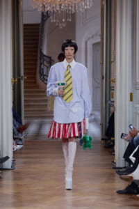 Model walks a grand hallway runway wearing an oversized light-blue shirt, yellow striped tie, red-white striped mini skirt, and white lace-up boots, holding a green plush accessory.