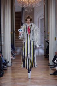 Model on a runway wearing a vertical-striped dress in yellow, gray, white with a chunky silver knitted jacket, holding a small white handheld device. A chandelier hangs above in an ornate hallway.