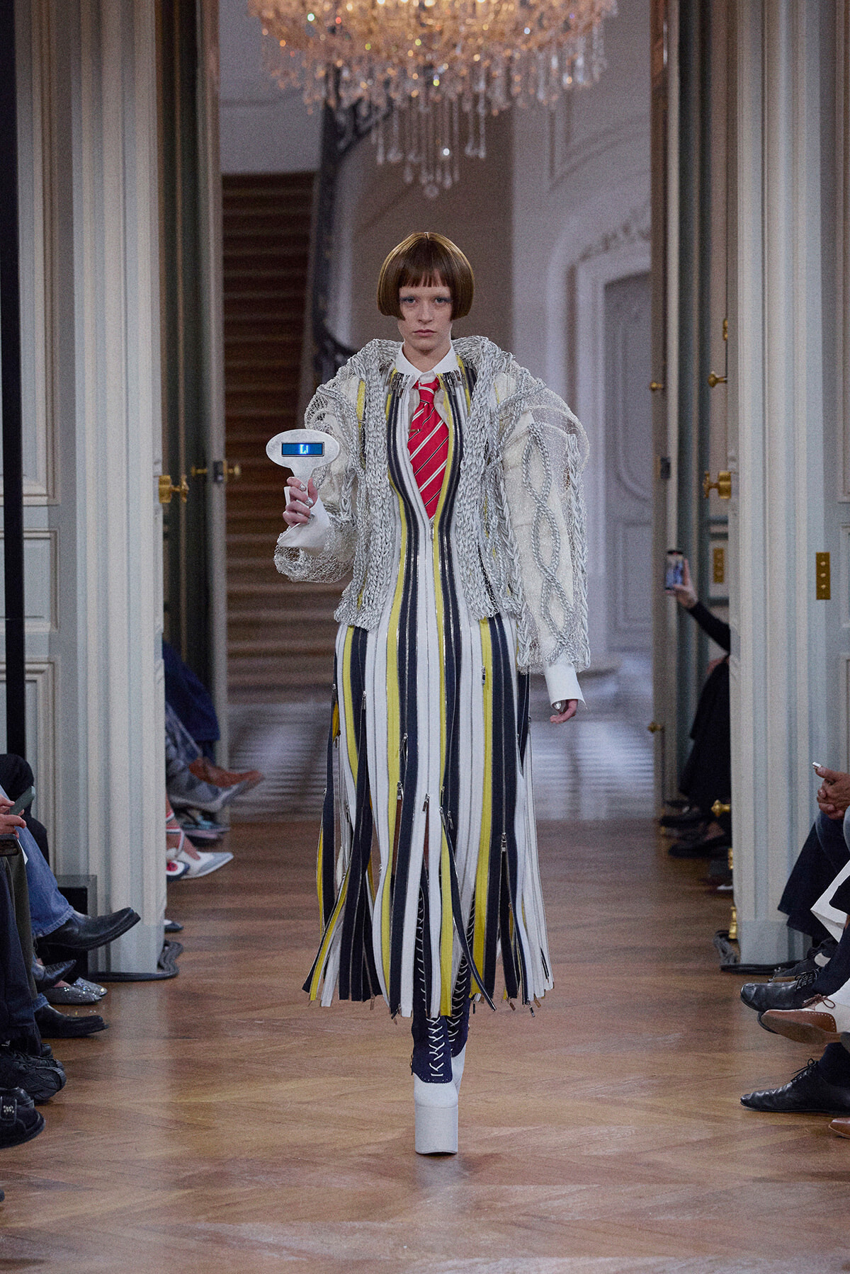 Model on a runway wearing a vertical-striped dress in yellow, gray, white with a chunky silver knitted jacket, holding a small white handheld device. A chandelier hangs above in an ornate hallway.