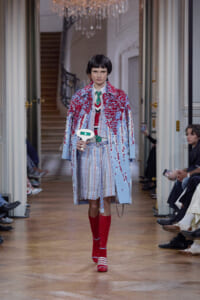 Model walks a runway in a pale blue coat splashed with red appliqués over a striped dress, paired with red knee-high socks and red-and-white striped shoes. She holds a small white prop.