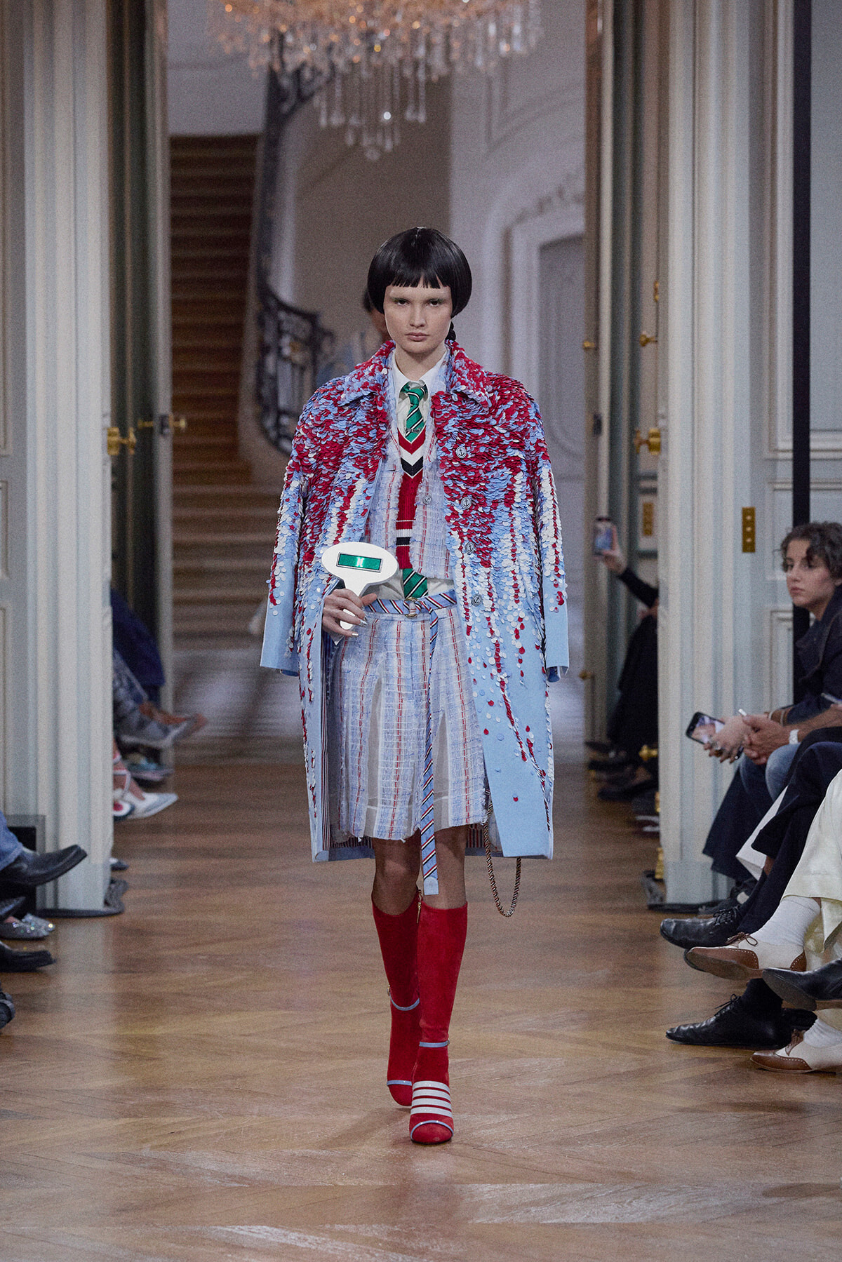 Model walks a runway in a pale blue coat splashed with red appliqués over a striped dress, paired with red knee-high socks and red-and-white striped shoes. She holds a small white prop.