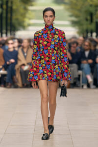 Runway model wearing a bold floral turtleneck mini dress in red, blue, and yellow, with a small black handbag and black shoes.