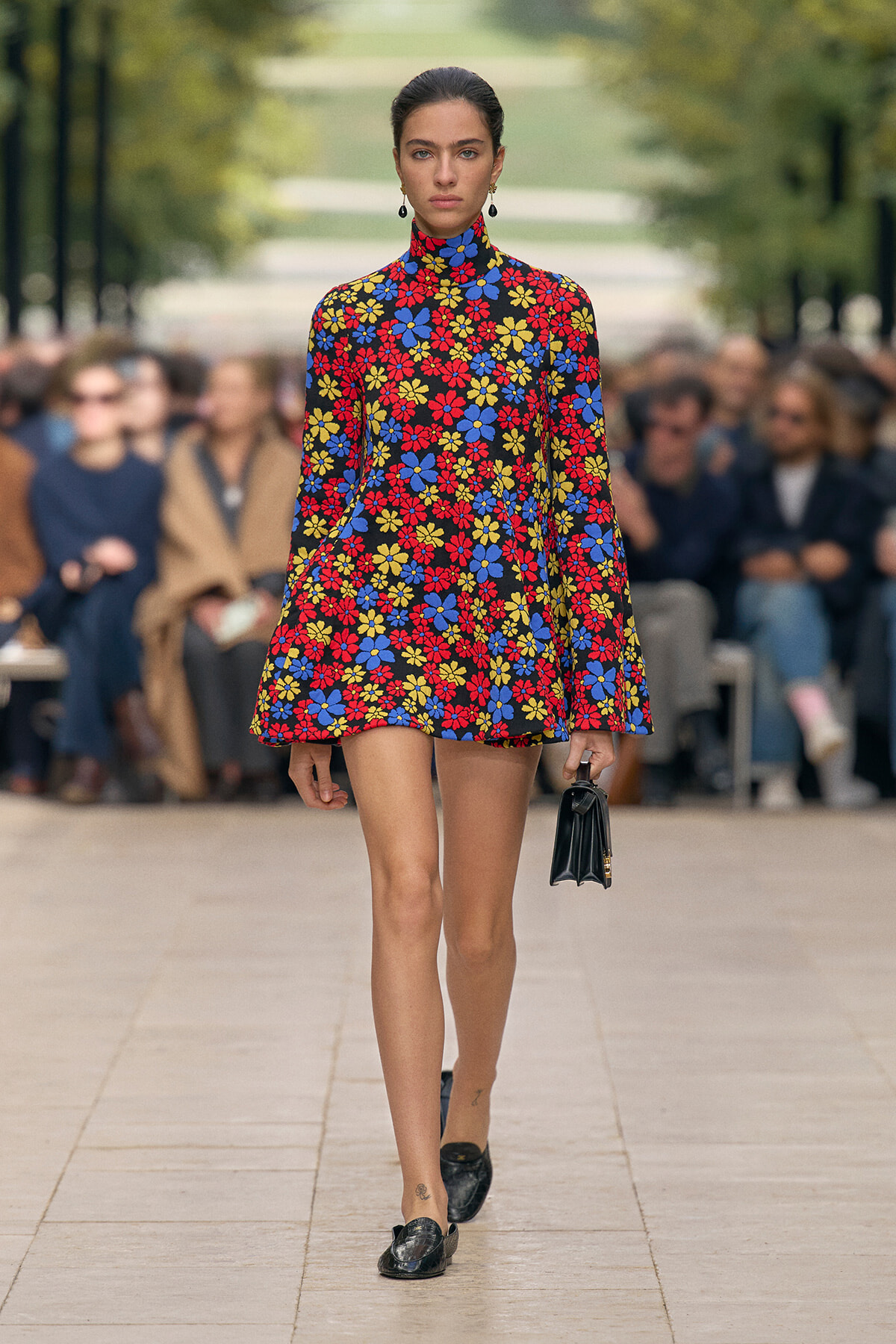 Runway model wearing a bold floral turtleneck mini dress in red, blue, and yellow, with a small black handbag and black shoes.