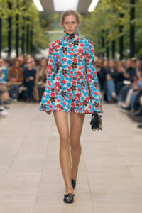 Runway model walks a catwalk in a blue, red, and brown floral turtleneck mini dress, carrying a black clutch.