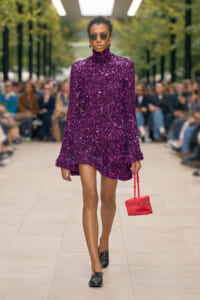 Model walking a runway in a purple sequined turtleneck mini dress, black shoes, round sunglasses, and a small red handbag; outdoor fashion show setting.