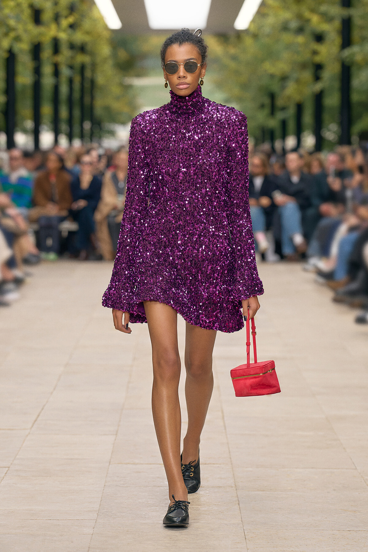 Model walking a runway in a purple sequined turtleneck mini dress, black shoes, round sunglasses, and a small red handbag; outdoor fashion show setting.