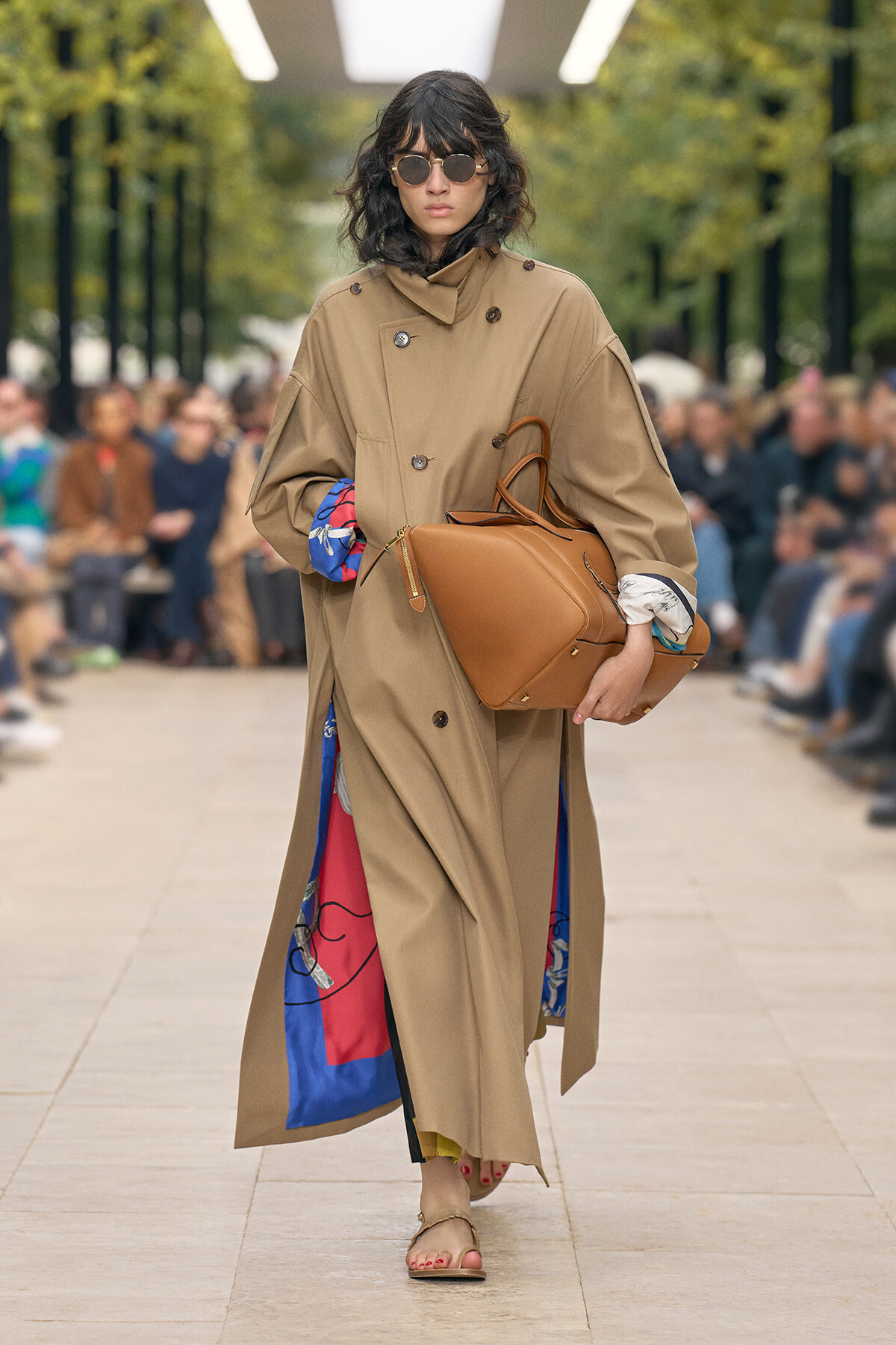 Model walking a runway in a beige oversized trench, round sunglasses, and carrying a large tan leather bag with colorful lining peeking out.