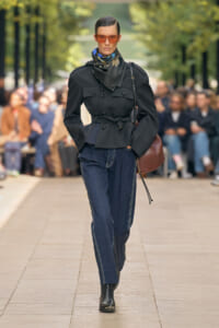 Model walks an outdoor runway in a black belted, double-breasted jacket, dark denim jeans, scarf, and orange sunglasses with a brown shoulder bag.