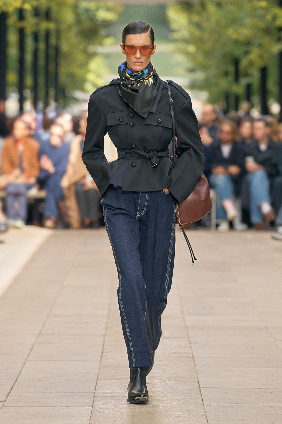 Model walks an outdoor runway in a black belted, double-breasted jacket, dark denim jeans, scarf, and orange sunglasses with a brown shoulder bag.