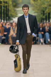 Male model in a dark blazer and brown trousers walks a runway, carrying a black helmet and a straw tote bag; audience blurred in background.