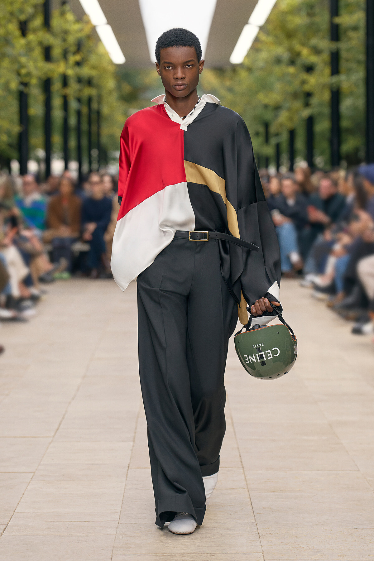 Male model on a fashion show runway wearing a color-block shirt in red, black, white and gold, with black wide-leg pants, holding a green helmet with a logo.