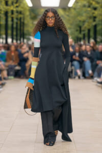 Model on a runway wearing a long black turtleneck dress with color-blocked sleeves, sunglasses, and a brown handbag.