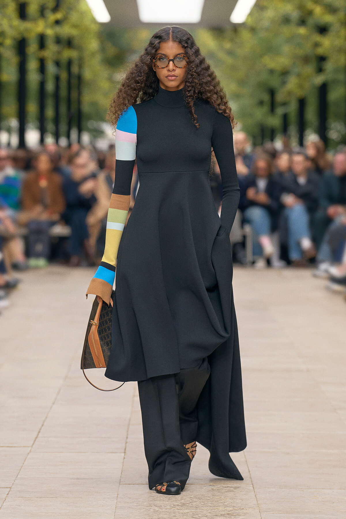 Model on a runway wearing a long black turtleneck dress with color-blocked sleeves, sunglasses, and a brown handbag.