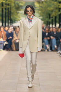 Model on a runway wearing an oversized cream blazer over white shirt and pants, with bold layered beaded necklaces, sunglasses, and a small red round clutch.