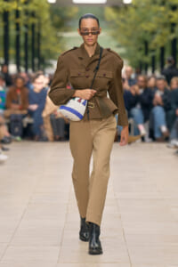 Model on a runway wearing a brown military-style jacket, tan pants, black ankle boots, sunglasses, and a white striped crossbody bag.