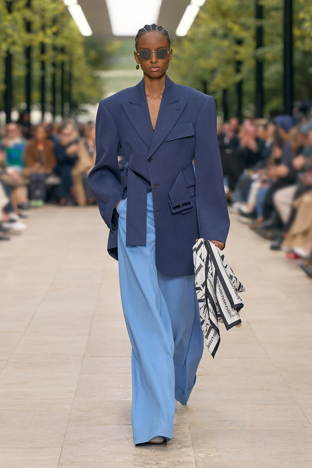 Model walks a fashion runway in an oversized navy blazer with large pockets and light-blue wide-leg trousers, wearing round sunglasses and holding a black-and-white patterned scarf.