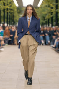 Model on a runway wearing an oversized navy blazer, light blue shirt, brown tie, and tan pleated trousers with black ankle boots.