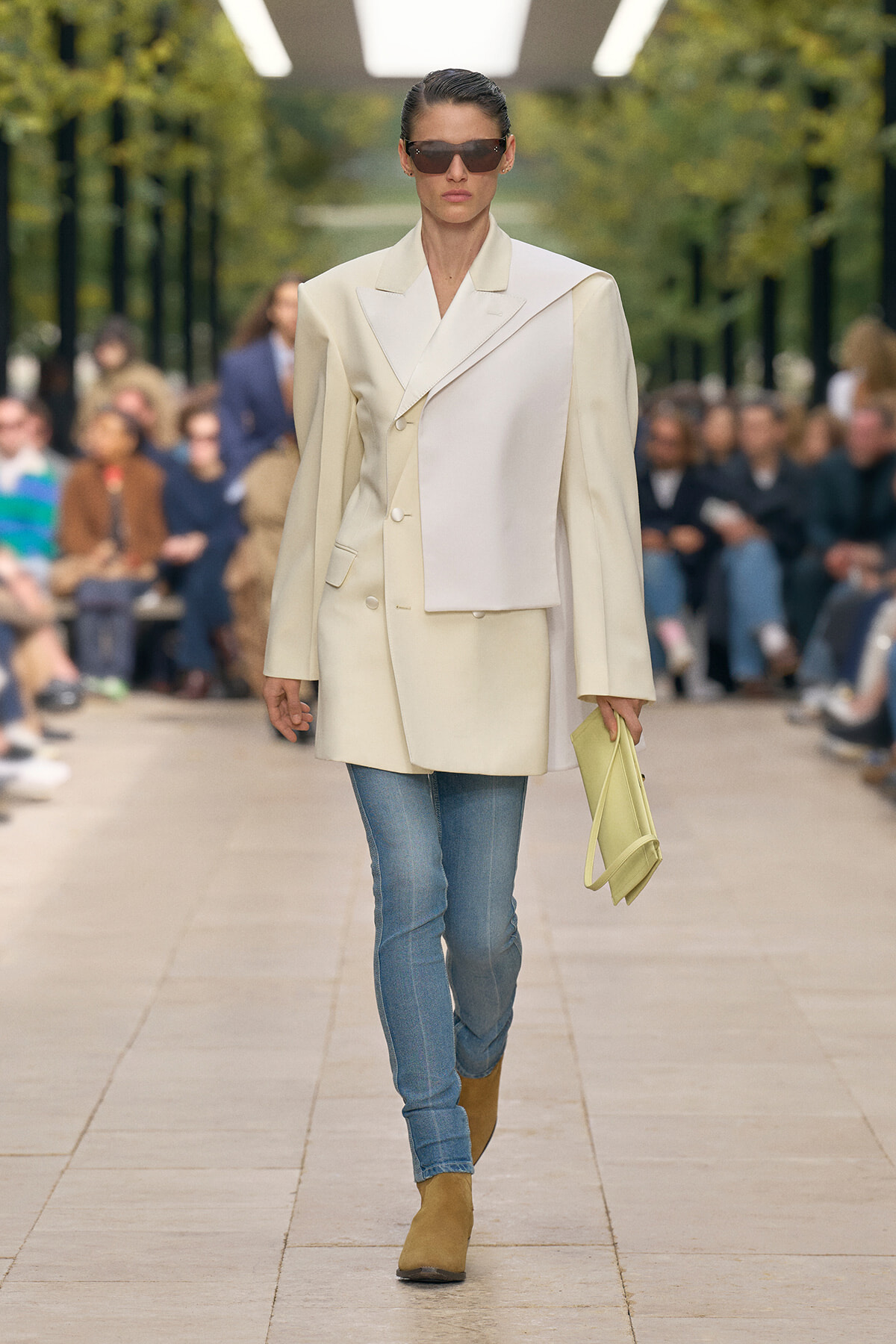 Model walking a runway in an oversized cream blazer with an asymmetric panel, paired with blue jeans, tan ankle boots, and large sunglasses, carrying a pale yellow clutch.