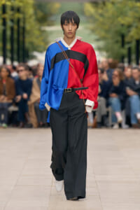 Model walks a fashion runway wearing a color-blocked red, blue, and black oversized blouse with wide-leg black pants.