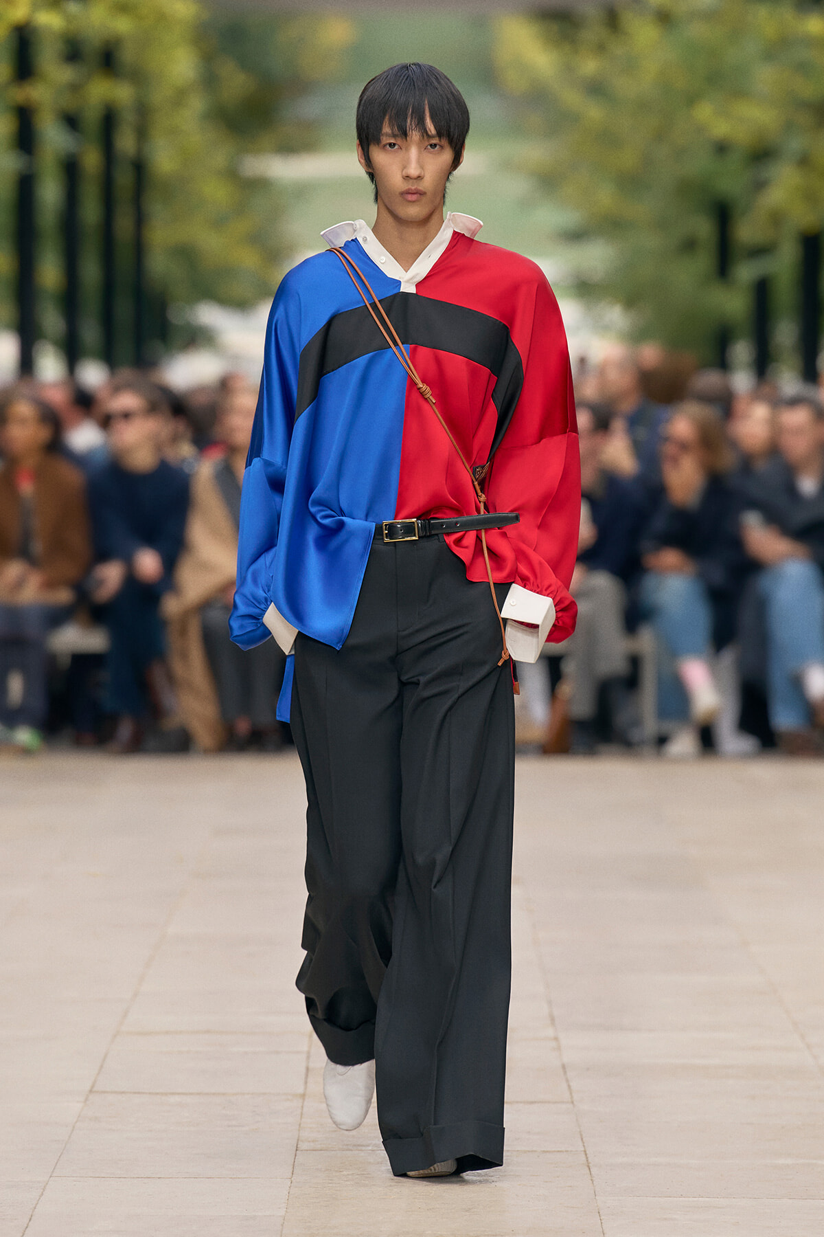 Model walks a fashion runway wearing a color-blocked red, blue, and black oversized blouse with wide-leg black pants.