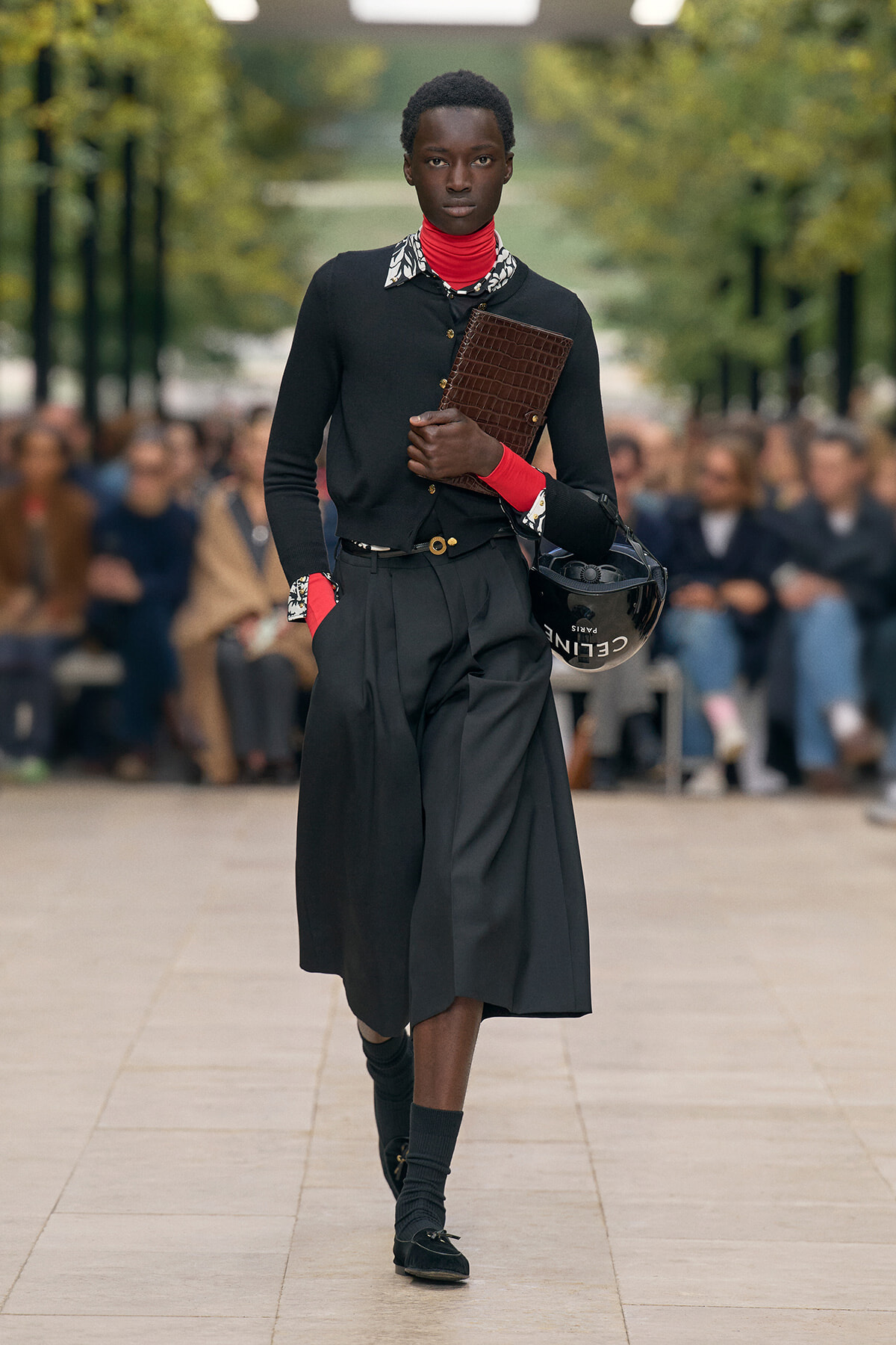 Model walks a runway in a black cardigan and wide-leg skirt, holding a brown textured clutch and a black helmet under arm.