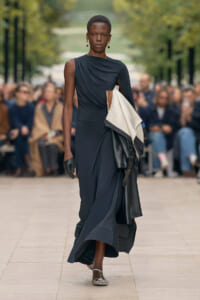 Model on a runway wearing a dark navy one-shoulder dress, carrying folded fabric swatches and leather pieces over her arm.