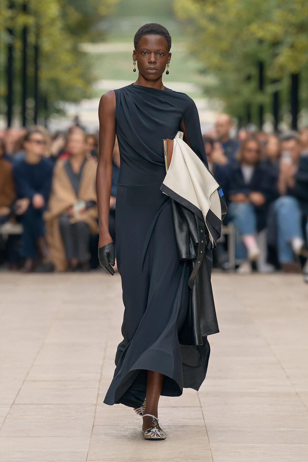 Model on a runway wearing a dark navy one-shoulder dress, carrying folded fabric swatches and leather pieces over her arm.
