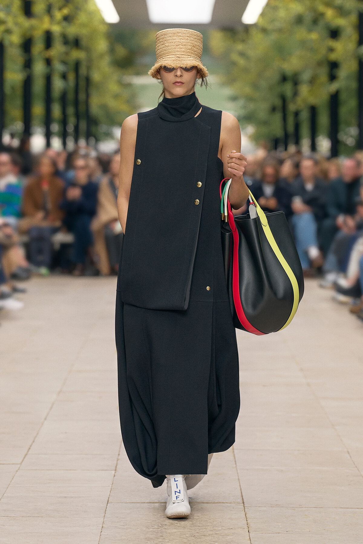 Model on a runway wearing an all-black sleeveless outfit and a straw hat, carrying a large black bag with colorful straps.