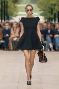 Model walking a runway in a short black textured dress with a scalloped neckline, sunglasses, and a brown handbag. Audience blurred in background.