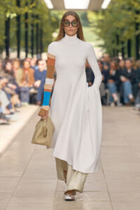Model walks a runway in a white turtleneck midi dress with a color-block sleeve, carrying a beige handbag and wearing oversized sunglasses.