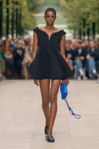 Model walking a runway in a short black textured dress with a deep V neckline, holding a bright blue clutch and wearing small, cat-eye glasses.