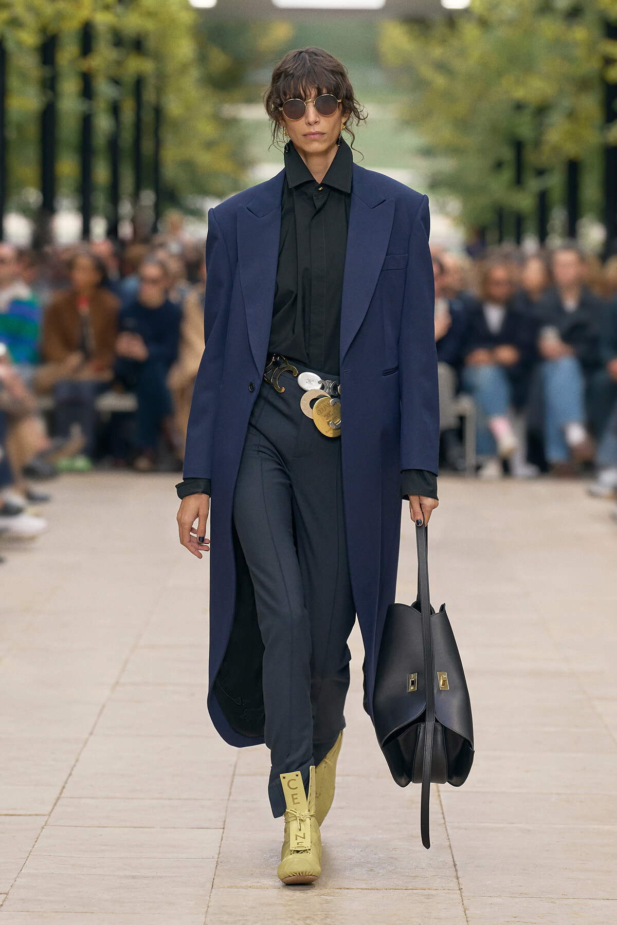 Model walks a runway in a long navy blue coat over a black shirt and dark trousers, accessorized with a wide belt and stacked metallic medallions, and carries a large black tote bag; pale yellow boots with text detail visible.