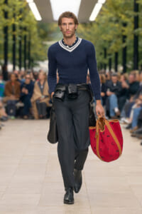 Male model walking a runway in a navy knit sweater, dark trousers, leather jacket tied at the waist, and a red tote bag with a pattern.