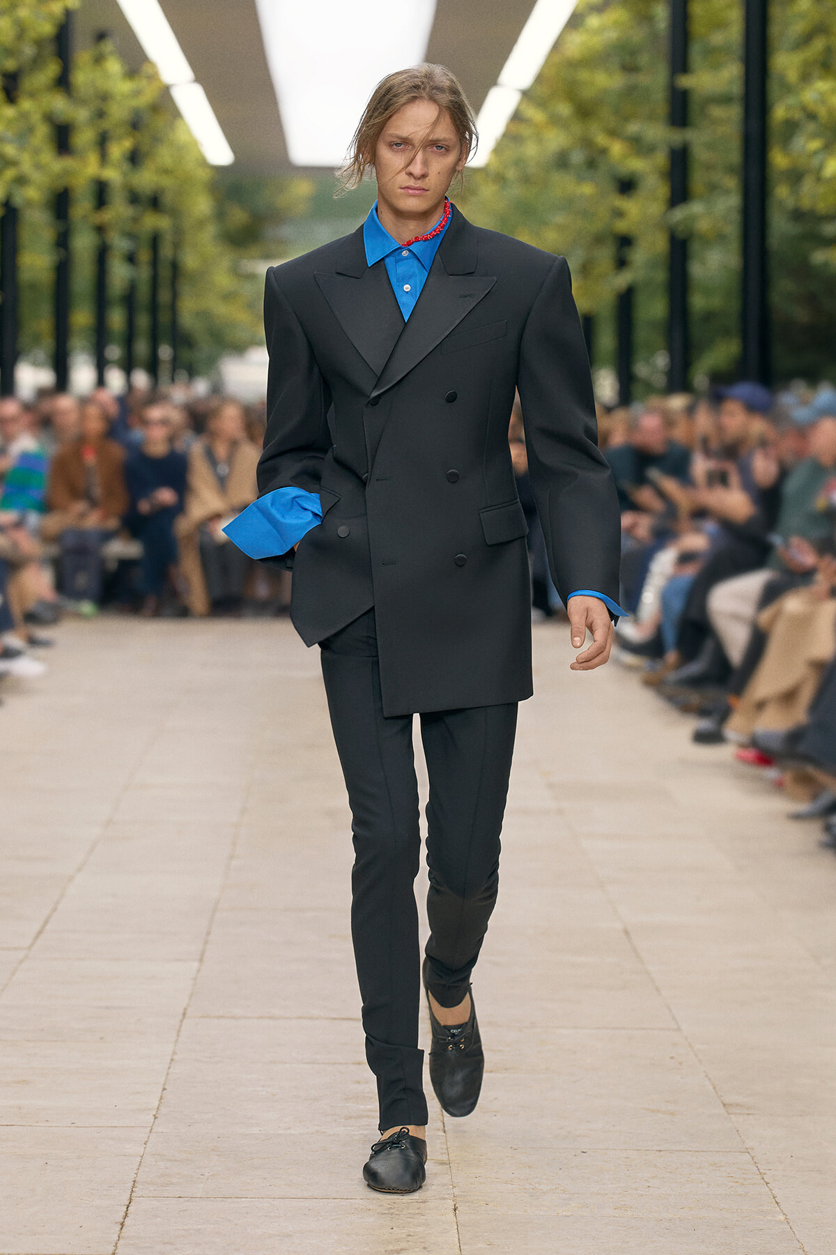 Male runway model wearing an oversized black double-breasted blazer with blue dress shirt sleeves and collar, walking down a fashion show runway.