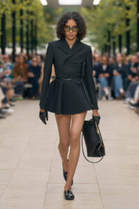 Runway model in a black double‑breasted blazer mini‑dress with a waist belt, gloves, sunglasses, and a black handbag.