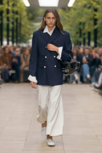 Model walks a runway in a navy double-breasted blazer with white shirt and wide white trousers, carrying a black helmet bag.