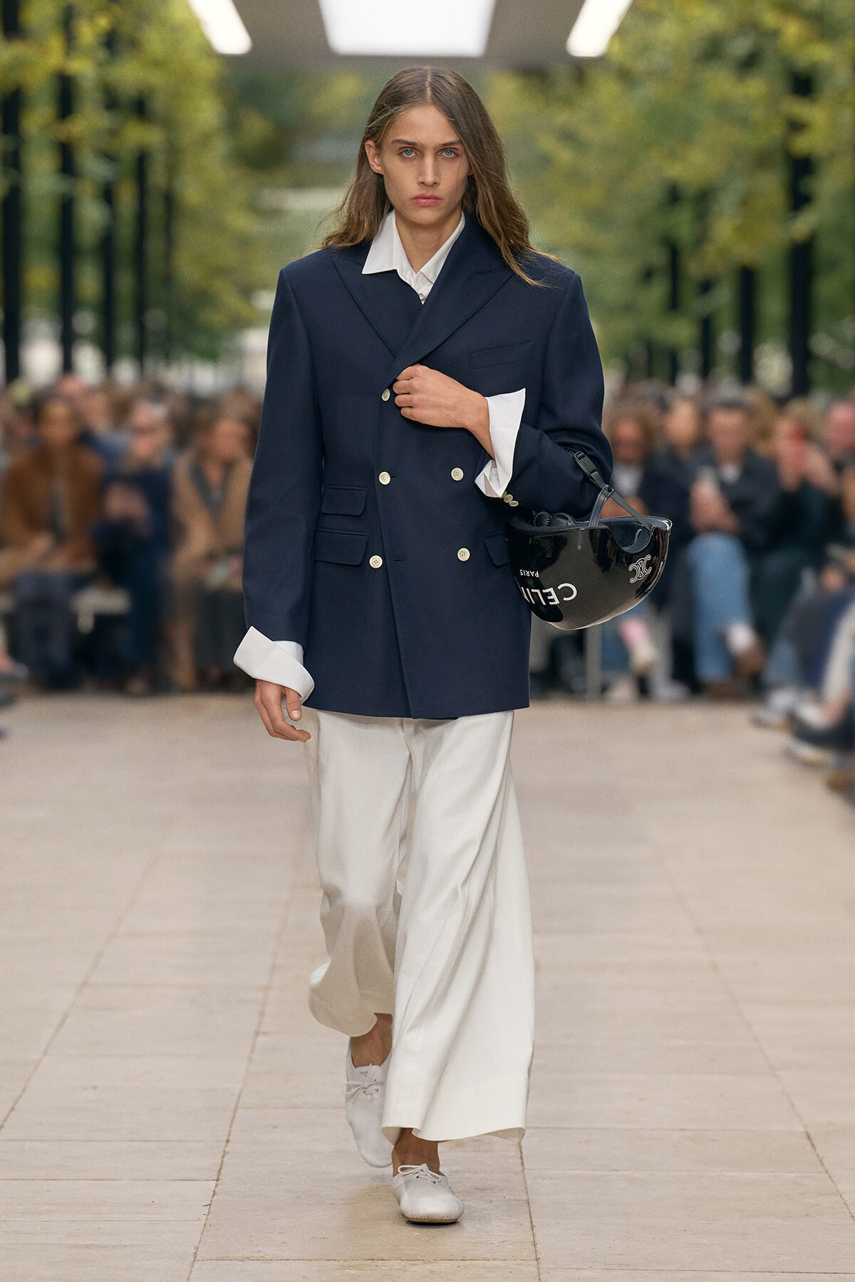 Model walks a runway in a navy double-breasted blazer with white shirt and wide white trousers, carrying a black helmet bag.